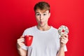 Young caucasian man drinking a cup of coffee and croissant making fish face with mouth and squinting eyes, crazy and comical