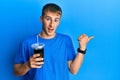 Young caucasian man drinking glass of soda beverage pointing thumb up to the side smiling happy with open mouth