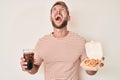 Young caucasian man eating fast food angry and mad screaming frustrated and furious, shouting with anger looking up Royalty Free Stock Photo