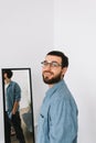 Young caucasian man in glasses standing at the mirror in living room