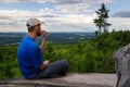 Young caucasian man, hiker in cap sitting and eating dinner, resting on rock and looking to valley. Czech summer landscape Royalty Free Stock Photo