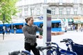 Young caucasian man hiring bicycle with machine in city, road trafic and pedestrians in background.