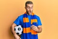 Young caucasian man holding football ball looking at smartphone afraid and shocked with surprise and amazed expression, fear and Royalty Free Stock Photo