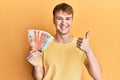 Young caucasian man holding philippine peso banknotes smiling happy and positive, thumb up doing excellent and approval sign Royalty Free Stock Photo