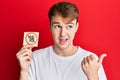 Young caucasian man holding under 18 prohibition sticker pointing thumb up to the side smiling happy with open mouth Royalty Free Stock Photo