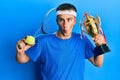 Young caucasian man playing tennis holding trophy making fish face with mouth and squinting eyes, crazy and comical