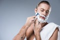 Young caucasian man with shaving foam on face holding razor in hand against his cheek, while bath towel laying on his shoulder, Royalty Free Stock Photo