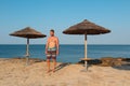 Young caucasian man in shorts, half body naked standing on Croatia sea shore edge with wicker umbrella. Front view