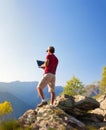 Young caucasian man sitting outdoor on a rock working on a laptop pc in mountain area. Royalty Free Stock Photo