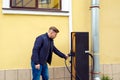 Young Caucasian man stands near electric car charging station and holds electrical connector to charge batteries. Lifestyle