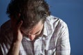 A young caucasian man with striped shirt sitting while looking down and holding his head with right hand against light blue Royalty Free Stock Photo