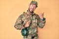 Young caucasian man wearing camouflage army uniform and canteen pointing thumb up to the side smiling happy with open mouth Royalty Free Stock Photo