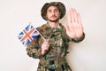 Young caucasian man wearing camouflage army uniform holding united kingdom flag with open hand doing stop sign with serious and Royalty Free Stock Photo