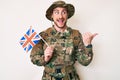 Young caucasian man wearing camouflage army uniform holding united kingdom flag pointing thumb up to the side smiling happy with Royalty Free Stock Photo