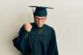 Young caucasian man wearing graduation cap and ceremony robe angry and mad raising fist frustrated and furious while shouting with Royalty Free Stock Photo