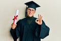 Young caucasian man wearing graduation cap and ceremony robe holding diploma doing ok sign with fingers, smiling friendly Royalty Free Stock Photo