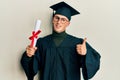 Young caucasian man wearing graduation cap and ceremony robe holding diploma smiling happy and positive, thumb up doing excellent Royalty Free Stock Photo