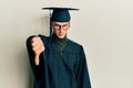 Young caucasian man wearing graduation cap and ceremony robe looking unhappy and angry showing rejection and negative with thumbs Royalty Free Stock Photo