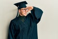 Young caucasian man wearing graduation cap and ceremony robe very happy and smiling looking far away with hand over head Royalty Free Stock Photo