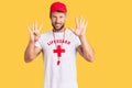 Young caucasian man wearing lifeguard t shirt holding whistle showing and pointing up with fingers number nine while smiling