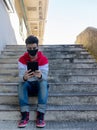 Young caucasian man working on his tablet, while sitting on some stairs, outdoors, with a face mask Royalty Free Stock Photo