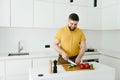 Young caucasian man in yellow t-shirt cooking healthy vegetable meal or salad in white modern kitchen with a knife smiling Royalty Free Stock Photo