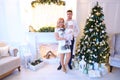 Young caucasian mother and father keeping little female baby near Christmas tree and decorated fireplace.