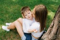 Young Caucasian mother kisses her three-year-old son on the cheek while sitting in the park near a tree on the grass in summer Royalty Free Stock Photo