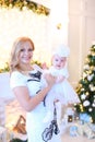 Young caucasian mother wearing white dress and keeping little female baby near Christmas tree.