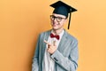 Young caucasian nerd man wearing glasses and graduation cap smiling cheerful pointing with hand and finger up to the side Royalty Free Stock Photo