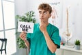 Young caucasian physio man holding call me banner at the clinic serious face thinking about question with hand on chin, thoughtful Royalty Free Stock Photo