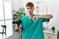 Young caucasian physio man holding muscle percusion gun at the clinic with angry face, negative sign showing dislike with thumbs