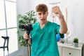 Young caucasian physio man holding muscle percusion gun at the clinic annoyed and frustrated shouting with anger, yelling crazy