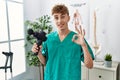 Young caucasian physio man holding muscle percusion gun at the clinic doing ok sign with fingers, smiling friendly gesturing