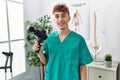 Young caucasian physio man holding muscle percusion gun at the clinic looking positive and happy standing and smiling with a