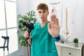 Young caucasian physio man holding muscle percusion gun at the clinic with open hand doing stop sign with serious and confident