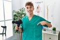 Young caucasian physio man holding muscle percusion gun at the clinic pointing finger to one self smiling happy and proud
