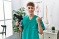 Young caucasian physio man holding muscle percusion gun at the clinic pointing thumb up to the side smiling happy with open mouth