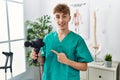 Young caucasian physio man holding muscle percusion gun at the clinic smiling happy pointing with hand and finger