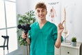 Young caucasian physio man holding muscle percusion gun at the clinic smiling happy pointing with hand and finger to the side