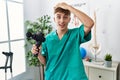 Young caucasian physio man holding muscle percusion gun at the clinic stressed and frustrated with hand on head, surprised and