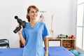 Young caucasian physio therapist girl smiling happy holding gun percusion at the clinic