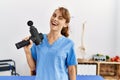 Young caucasian physio therapist girl smiling happy holding gun percusion at the clinic