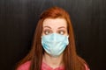 Young crazy tired redhaired girl with beveled eyes in blue mask, pink sweater on black background, coronavirus epidemic