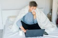 Young caucasian sick man blowing his nose sitting on the bed under a warm blanket and working on a laptop. Work at home during an Royalty Free Stock Photo