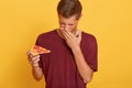 Young Caucasian student male fast food lover dresses burgundy t shirt gets his order from pizzeria, holding slice of expired pizza Royalty Free Stock Photo