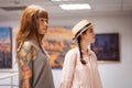 Young Caucasian tanned woman with tattoo and girl wearing straw hat contemplating arts. In background is exhibition hall