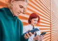 Young caucasian teens girl and boy portrait. Teenagers browsing smartphone devices standing near orange wall. Careless young