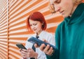 Young caucasian teens girl and boy portrait. Teenagers browsing smartphone devices standing near orange wall. Careless young