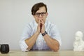 Young caucasian very serious male in white robe sits frowning, hands on the table. Feeling look, strong emotions. Electronic watch